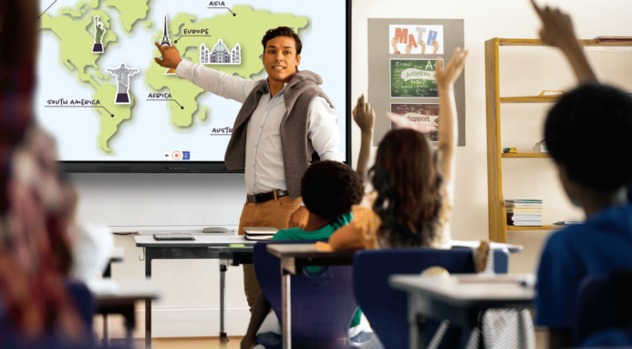A teacher stands in front of a classroom using a Promethean board while students raise their hands, ready to ask questions.