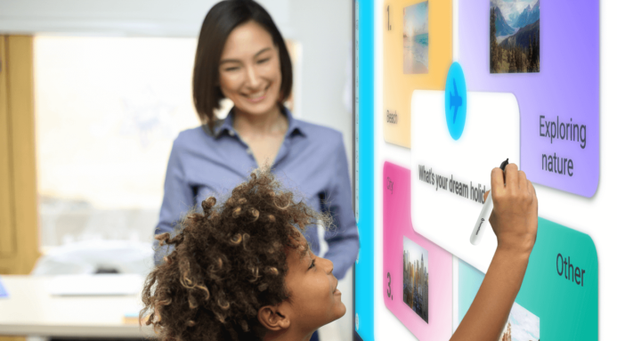 A young schoolgirl is using an interactive whiteboard while her teacher observes with an eager smile.