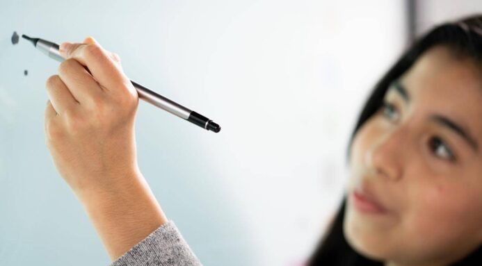 A school child stands in front of an interactive whiteboard, holding a stylus in their hand, concentrating on the start of a drawing.